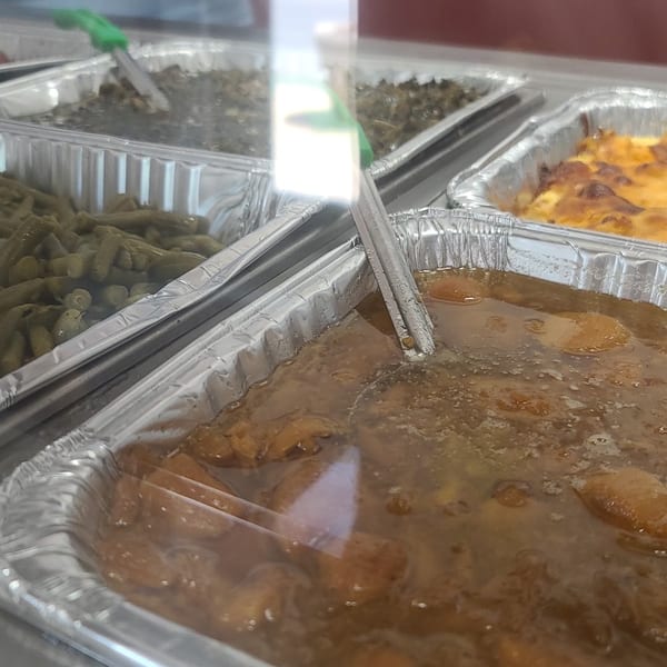 Buffet line with trays of green beans, collard greens, macaroni and cheese, and stewed chicken.