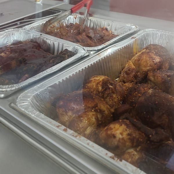 Three foil pans of cooked meats, including ribs with sauce and seasoned chicken pieces, in a display case.